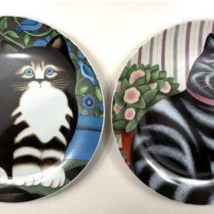 May include: Two round ceramic plates featuring cat illustrations. The left plate shows a black and white cat amidst blue flowers and birds. The right plate displays a grey and black striped cat with a pink collar, sitting near a potted plant.