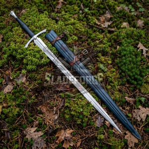 May include: A long, silver sword with a black handle and crossguard, resting on a bed of green moss and fallen leaves. The sword is next to a blue sheath with brown leather straps. The text "SLADEFORGE" is partially visible.