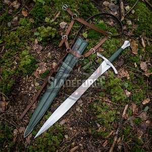 Puede incluir: Una espada medieval con una hoja plateada y un mango verde, sobre musgo y hojas. La espada está acompañada de una vaina verde y correas de cuero marrón. La imagen está tomada desde una perspectiva cenital.