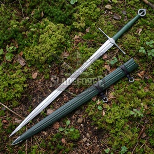 Puede incluir: Una espada larga y plateada con empuñadura y guarda verdes, sobre un lecho de musgo verde. La espada va acompañada de una vaina verde a juego. La imagen está tomada desde un ángulo ligeramente elevado.