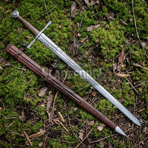 Peut inclure: Une longue épée argentée avec une poignée et une garde transversale marron repose sur de la mousse verte. Un fourreau en cuir marron est posé à côté. La lame de l'épée est gravée d'un motif et la poignée a un pommeau arrondi.