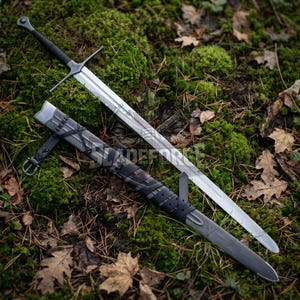 Op de afbeelding: Een lang, zilveren zwaard met een zwart handvat en pareerstang, liggend op een bed van groen mos en gevallen bladeren. De schede van het zwaard is zwart en grijs met leren riemen. Het lemmet is ongeveer 91 cm lang.