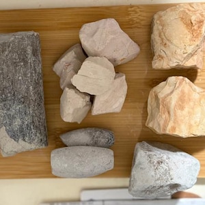 May include: An assortment of rocks and stones in shades of grey, beige, and reddish-brown, displayed on a wooden chopping board. The collection features a variety of textures, including smooth and rough surfaces.