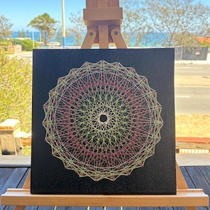 Puede incluir: Arte de hilo geométrico sobre lienzo negro, con un diseño circular de hilos amarillos, rosas y verdes. La obra se exhibe en un pequeño caballete de madera, con un fondo costero visible.