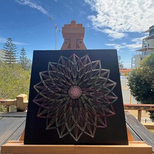 May include: A black square string art piece featuring a geometric floral design in pink, green, and yellow threads. The artwork is displayed on a wooden easel, set against a backdrop of a blue sky and clouds.