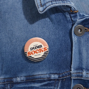 Puede incluir: Un botón redondo con el texto "Andy's DUMB SOCKS Estd 2026" en rojo, blanco y azul. El botón está sobre una chaqueta vaquera azul.