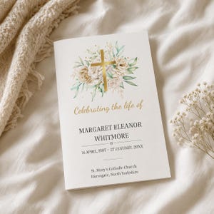 May include: A white funeral program booklet featuring a gold cross surrounded by watercolour roses and greenery. The text reads: Celebrating the life of MARGARET ELEANOR WHITMORE, 14 APRIL, 1997 – 27 JANUARY, 20XX, St. Mary's Catholic Church, Harrogate, North Yorkshire.