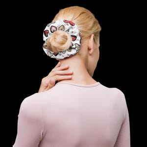 May include: A white scrunchie with a pattern of red, gold, and gray hearts. The scrunchie is wrapped around a bun hairstyle. The person is wearing a light pink long-sleeved shirt.