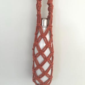 May include: A white metal water bottle encased in a rust-coloured macrame holder featuring a diamond-shaped knotted pattern. The holder includes a long, braided handle with two light-coloured wooden rings at the top for hanging or carrying.