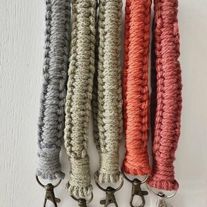 May include: Five macrame wristlet lanyards arranged vertically. The lanyards feature braided cord in grey, sage green, beige, burnt orange, and dusty rose. Each lanyard is finished with a metal lobster clasp at the bottom end for attachment.