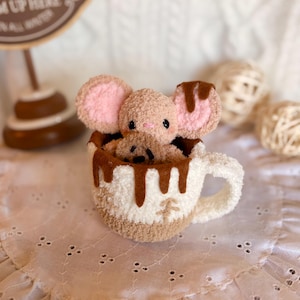 Könnte beinhalten: Eine beigefarbene Plüschmaus mit rosa Innenohren sitzt in einer gehäkelten Tasse in Weiß und Beige. Die Tasse ist am Rand mit braunen Filztropfen verziert, die Schokolade imitieren, und hat ein kleines braunes Stickdetail auf der Vorderseite.