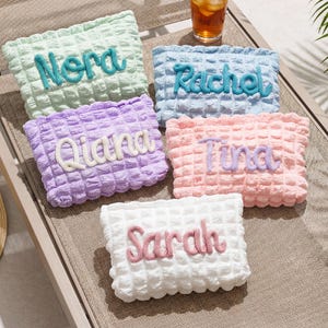 May include: Five pastel-coloured cosmetic bags with textured surfaces, each embroidered with a name. The colours are light green, light blue, lavender, pink, and white. The names embroidered are Nora, Rachel, Qiana, Tina, and Sarah. The bags are on a sun lounger.