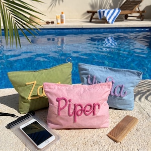 May include: Three personalized cosmetic bags in green, pink, and blue, each with a name embroidered in a contrasting color. A phone in a waterproof case and a wooden comb sit nearby, with a pool and lounge chair in the background.