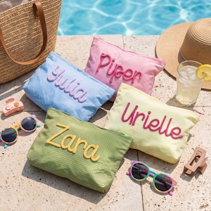 May include: A collection of personalized cosmetic bags in pastel colors: light blue, pink, yellow, and green. Each bag features a name embroidered in a contrasting color. Accessories include sunglasses, a straw hat, and a tote bag, suggesting a summer theme.