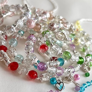 May include: A selection of beaded bracelets featuring a mix of colourful beads, including red, purple, green, and clear crystal beads. Some bracelets have silver accents and heart-shaped charms. The bracelets are displayed on a white surface.