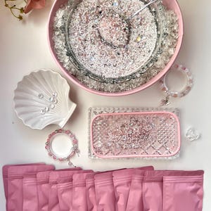 Könnte beinhalten: Draufsicht auf Bastelbedarf. Eine rosa Schüssel und ein rechteckiger Tablett sind mit kleinen, weißen und rosa Perlen gefüllt. Mehrere rosa Beutel sind unten angeordnet. Eine Muschelschale und zwei Armbänder sind ebenfalls zu sehen.