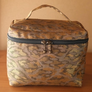 May include: A patterned cosmetic bag with a handle. The bag has a repeating design in gold, brown, and grey. It features a black zip and two decorative pulls. The bag is on a wooden surface.