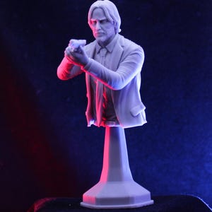 May include: A detailed, light gray bust of a man in a suit, holding a gun. The sculpture is mounted on a pedestal. The figure has a beard and long hair. The background is dark with red and blue lighting.