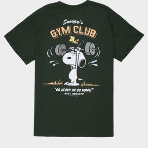 May include: Dark green t-shirt featuring Snoopy lifting weights with Woodstock. The shirt has the text "Snoopy's GYM CLUB" and "GO HEAVY OR GO HOME!" printed on it. The brand name "RIOT SOCIETY" is also printed on the shirt.
