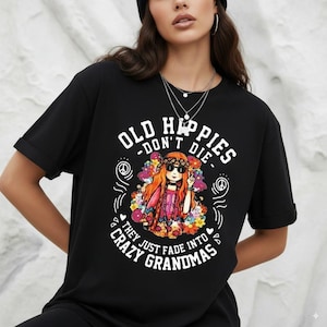 May include: Black t-shirt with the text "OLD HIPPIES - DON'T DIE - THEY JUST FADE INTO CRAZY GRANDMAS." The graphic features a cartoon woman with sunglasses, flowers, and a peace sign. The shirt is a casual style.