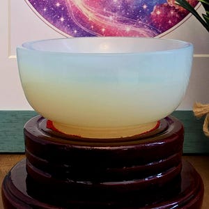 May include: A translucent opalescent glass bowl with a smooth, rounded rim and a soft, milky white to pale yellow gradient appearance. The bowl is positioned securely on top of a dark, polished, multi-tiered circular wooden display stand.