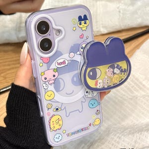 Op de afbeelding: Een doorzichtige telefoonhoes met een paarse tint, versierd met cartoonfiguren en het woord "Love". Een bijpassende paarse en gele telefoonhouder is bevestigd. De hoes is ontworpen voor een telefoon met twee cameralenzen.