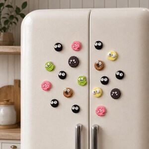 May include: A collection of small, round refrigerator magnets shaped like cookies and circular treats in various colors including black, pink, green, yellow, and brown. Each piece features two white plastic googly eyes attached to the surface.