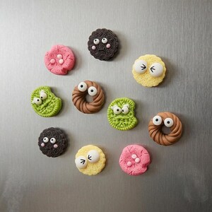 May include: A collection of ten round cookie-shaped magnets featuring various colors including pink, dark brown, yellow, and green. Each magnet has two white plastic eyes attached to the surface, with some showing small pink cheeks or black line details.