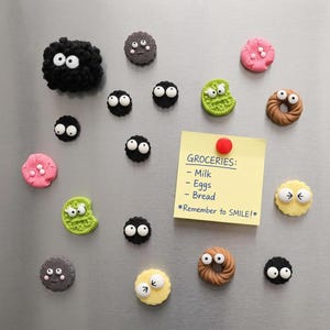 May include: A collection of decorative magnets featuring black fuzzy soot sprites, colorful textured cookies, and donut shapes with googly eyes. A yellow square sticky note with the text GROCERIES: Milk, Eggs, Bread, *Remember to SMILE!* is attached.