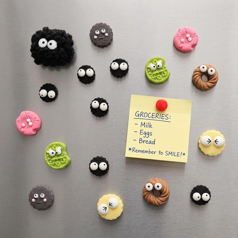 May include: A collection of decorative magnets featuring black fuzzy soot sprites, colorful textured cookies, and donut shapes with googly eyes. A yellow square sticky note with the text GROCERIES: Milk, Eggs, Bread, *Remember to SMILE!* is attached.
