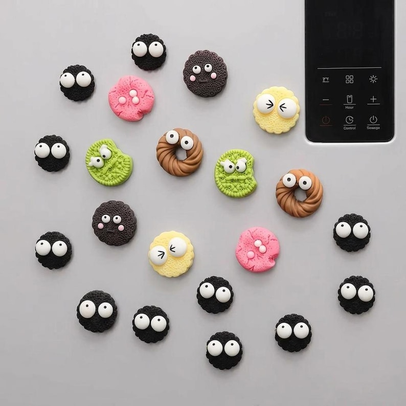 May include: A collection of twenty-one small, round, and ring-shaped magnets featuring cartoonish white eyes. The magnets are shaped like various cookies and soot sprites in colors including black, pink, yellow, green, and brown, arranged on a surface.