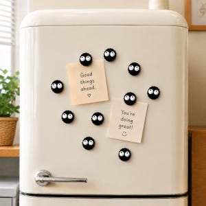 May include: A set of nine black, circular magnets with large white eyes and black pupils attached to a cream-colored refrigerator. Two square, light-colored sticky notes are held in place by the magnets, featuring the text Good things ahead and You're doing great.