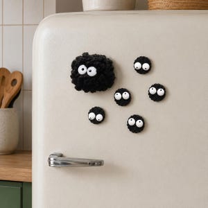 May include: A set of six black fuzzy circular magnets with large white googly eyes attached. One magnet is significantly larger and more textured than the other five smaller, flat, circular magnets. These decorative items are arranged on a cream refrigerator.