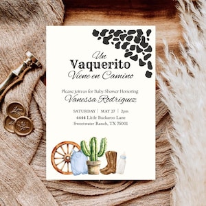 May include: A white baby shower invitation card featuring black cow print patterns, a wooden wagon wheel, a green cactus in a pot, a blue baby romper, a brown cowboy boot, and a baby bottle. The text reads Un Vaquerito Viene en Camino, Vanessa Rodriguez, Saturday May 27 2pm, 4444 Little Buckaroo Lane, Sweetwater Ranch, TX 75001.