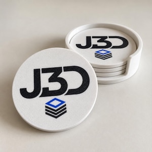 May include: A set of round white coasters with a textured surface. Each coaster features the black text J3D and a graphic icon consisting of three stacked black horizontal bars topped with a blue diamond shape. One coaster sits in front.