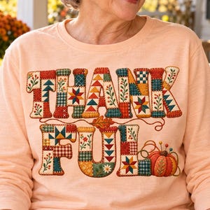 May include: A long-sleeve peach-colored shirt featuring the word THANKFUL printed in a patchwork quilt pattern. The letters are decorated with various geometric shapes, floral motifs, and a small orange pumpkin illustration at the end of the word.
