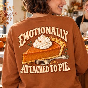 Puede incluir: Una camiseta de manga larga color óxido con un gráfico en la espalda de un trozo de tarta de calabaza con nata montada y un tenedor de plata. El texto sobre y bajo la tarta dice: "EMOTIONALLY ATTACHED TO PIE."