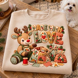 Può includere: Una felpa beige con la grafica di una renna che cuce una borsa patchwork contenente un orsacchiotto. Il testo "MADE WITH LOVE" appare sopra in un carattere trapuntato. Il design include motivi a quadretti rossi e verdi e dettagli floreali.