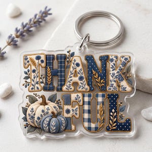 Op de afbeelding: Een doorzichtige acryl sleutelhanger met het woord THANKFUL in blauwe en crèmekleurige patronen, waaronder ruiten, stippen en bloemen. Twee pompoenen, één crème en één blauw, zijn onder de tekst geplaatst met enkele tarwehalmen.