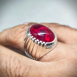 May include: A silver ring featuring a large, oval-shaped, deep red gemstone. The ring has a textured, ridged design on the sides and small decorative accents around the gemstone setting. The ring is shown on a person's finger.