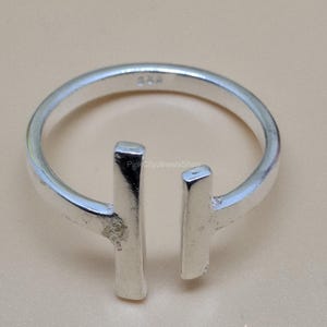 May include: A silver ring with a minimalist design. The ring features a band with two vertical rectangular bars that do not connect, creating an open design. The ring is set against a neutral background.