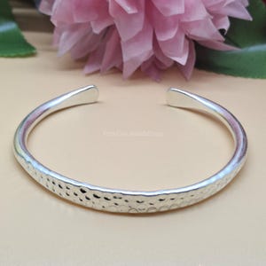 Puede incluir: Un brazalete de puño plateado martillado con una superficie texturizada y un diseño abierto. El fondo incluye una flor rosa y el texto "PinkCityJewelsShop".