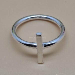 May include: A silver ring with a minimalist design. The ring features a thin band and a vertical rectangular bar at the center. The ring is set against a neutral background.