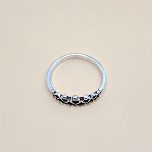Puede incluir: Un anillo de metal plateado con un diseño en forma de corona. La parte superior del anillo muestra una serie de pequeñas formas de corazón en relieve dispuestas en fila, acentuadas con detalles oscuros oxidados para resaltar el patrón.