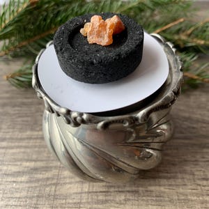 May include: A silver-colored incense burner with a decorative, swirling design. A black charcoal disc holds a piece of amber-colored resin. The burner sits on a wooden surface, with evergreen branches in the background.
