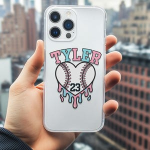 Puede incluir: Funda transparente para teléfono con un diseño temático de béisbol. El diseño presenta el nombre "TYLER" en rosa y azul, un corazón en forma de béisbol y el número "23" con acentos goteantes.
