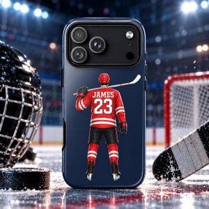 May include: A dark blue smartphone case featuring a graphic of a hockey player seen from behind, wearing a red jersey with the name JAMES and the number 23 in white. The player holds a hockey stick across their shoulders on the back of the case.
