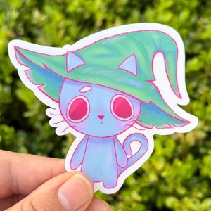 May include: A whimsical sticker featuring a blue cat wearing a green witch hat. The cat has large pink eyes and a matching pink outline. The sticker is held against a blurred green background, highlighting the vibrant colors and playful design.