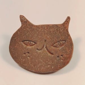 以下が含まれることがあります： 茶色の陶器製猫型ブローチ。表面はざらざらとした斑点模様で、アーモンド形の目、小さな鼻、両頬に2つの小さな点があります。耳は尖っており、全体的に丸みを帯びた平らな形状で、猫の顔を模したデザインになっています。