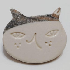 以下が含まれることがあります： 滑らかなオフホワイト仕上げの猫型セラミックブローチ。頭頂部はダークブラウンとグレーのまだら模様になっています。顔にはアーモンド型の目、シンプルな鼻、両頬に3つの小さな点が刻印されたディテールが施されています。